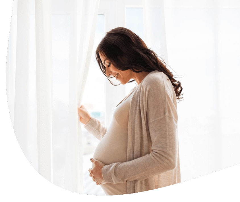 Usmívající se těhotná žena stojí u okna, drží se za břicho.