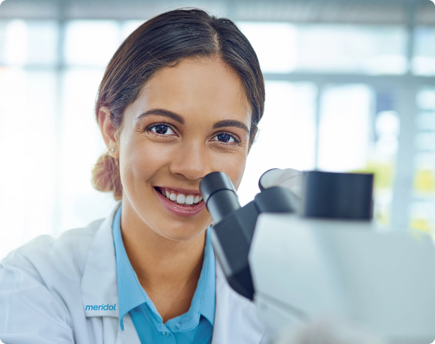 Usmívající se vědecká pracovnice u mikroskopu s logem „meridol" na svém laboratorním plášti, výjev symbolizuje zubní výzkum.