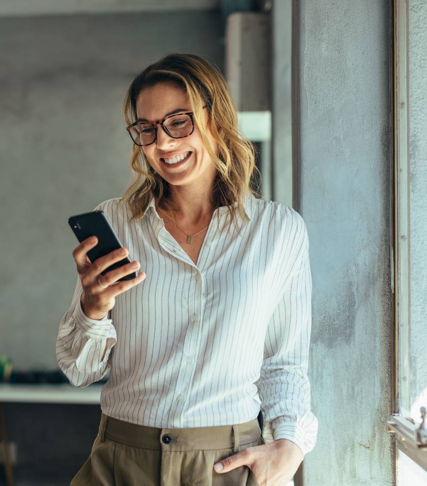 Šťastná žena s brýlemi čte zprávy na smartphonu ve světlé kanceláři.